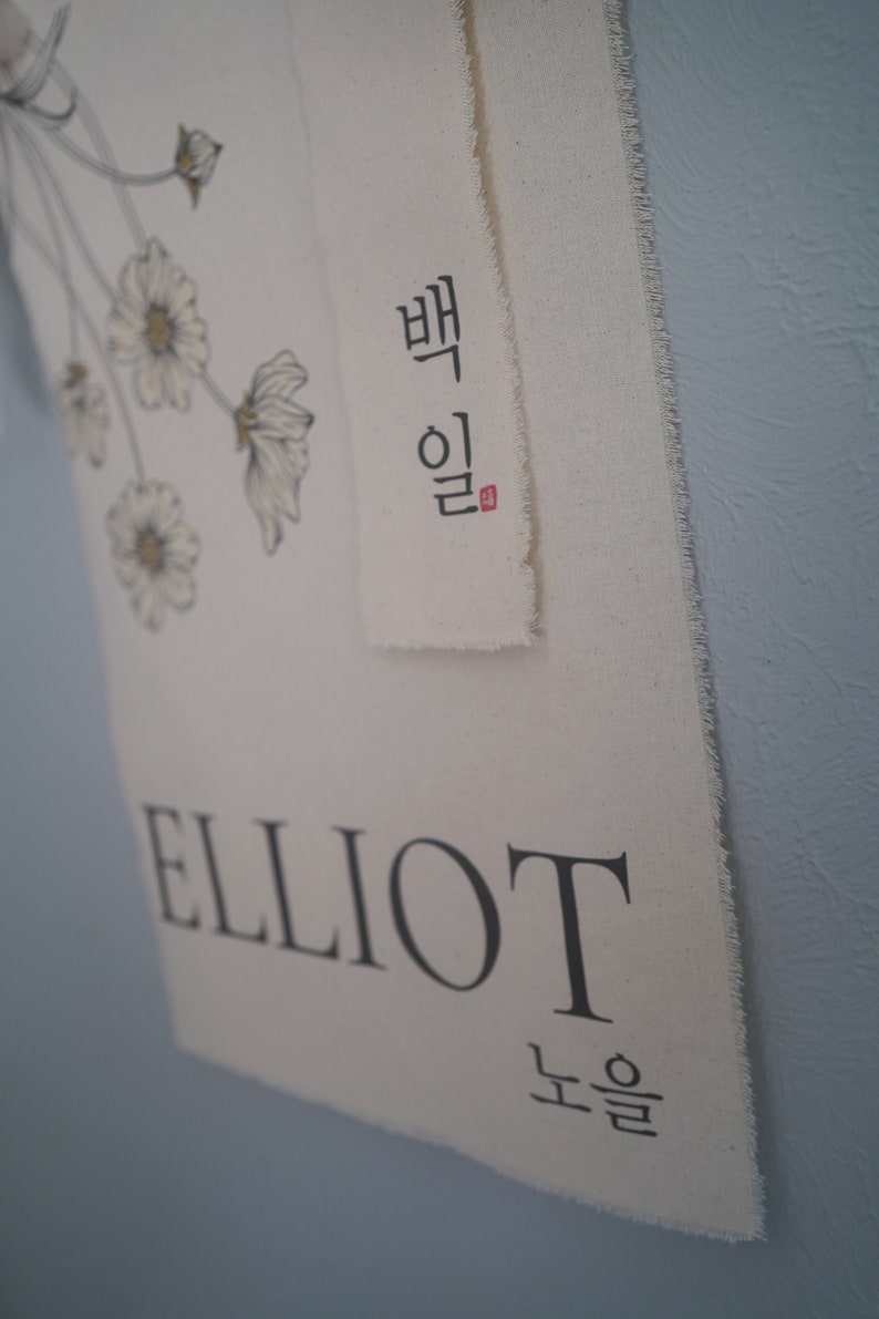 May include: A white canvas banner with the name "ELLIOT" printed in black. Above the name is a smaller banner with the Korean word "Elliot" written in black with a red seal. The banner is decorated with a floral design.