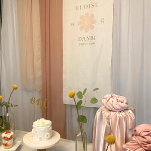 May include: A pink and white banner with the text "Eloise" and "Danbi Sweet Rain" with a pink flower graphic. The banner is hanging on a wall with pink and white fabric drapes. There is a white cake with pink and white flowers on a wooden cake stand. There are two glass vases with yellow flowers and green leaves. There are two pink gift boxes wrapped in satin fabric with yellow tassels.