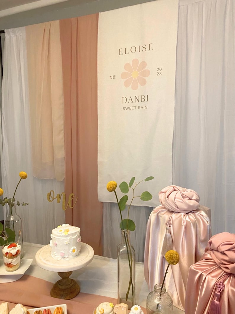 May include: A pink and white banner with the text "Eloise" and "Danbi Sweet Rain" with a pink flower graphic. The banner is hanging on a wall with pink and white fabric drapes. There is a white cake with pink and white flowers on a wooden cake stand. There are two glass vases with yellow flowers and green leaves. There are two pink gift boxes wrapped in satin fabric with yellow tassels.