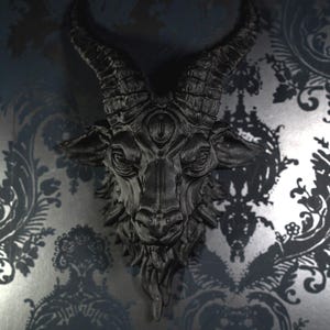 May include: A matte black wall-mounted sculpture of a goat head featuring prominent curved horns, a third eye centered on the forehead, and detailed facial features including a textured beard and pointed ears, presented in a monochromatic finish.