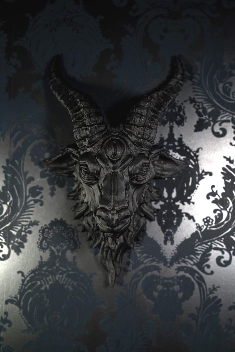May include: A matte black wall-mounted sculpture of a goat head featuring prominent curved horns, a third eye centered on the forehead, and detailed facial features including a textured beard and pointed ears, presented in a monochromatic finish.