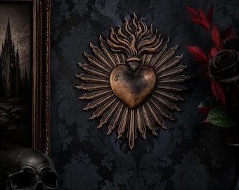 May include: A bronze-colored wall ornament shaped like a sacred heart with radiating sunburst rays and flames at the top. The heart features a small, teardrop-shaped indentation on its surface, finished with a dark, weathered metallic patina.