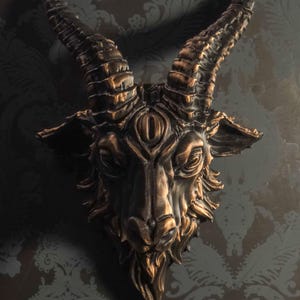 May include: A wall-mounted sculpture of a goat head featuring a third eye centered on the forehead. The piece is finished in a dark black tone with metallic bronze highlights accentuating the textured fur, curved horns, and facial features.