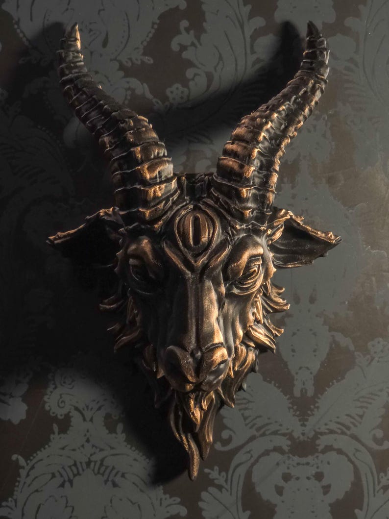 May include: A wall-mounted sculpture of a goat head featuring a third eye centered on the forehead. The piece is finished in a dark black tone with metallic bronze highlights accentuating the textured fur, curved horns, and facial features.