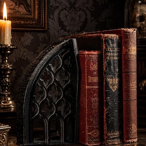 May include: A black metal bookend featuring an arched gothic window design with quatrefoil cutouts, positioned next to three antique hardcover books. The books include titles labelled The Demons, The College Standard Dictionary, and Dickens.