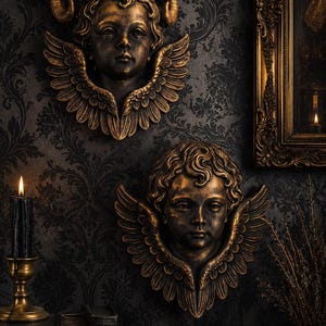 May include: Two wall-mounted sculptures of cherubic faces with feathered wings, finished in a metallic bronze tone. The upper sculpture features curved ram horns, while the lower sculpture displays curly hair. Both figures have detailed facial features.