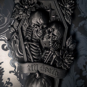 May include: A black wall plaque featuring a coffin shape with two skeletons facing each other. The design includes floral accents, a banner with the text "till death" embossed on it, a carved pumpkin, and a bat at the base, all in a matte black finish.