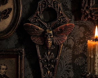 May include: A wall-mounted decorative plaque featuring a central human skull integrated with moth wings, finished in a dark bronze and black patina. The elongated, pointed design includes intricate scrollwork and a circular cutout at the top.