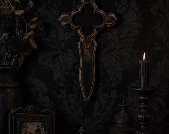 May include: A wall-mounted cross with a decorative, ornate frame featuring a dark, weathered finish. The cross has a central quatrefoil-shaped opening and a long, tapered lower section, crafted with a textured, aged appearance in shades of black and bronze.