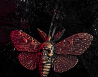May include: A bronze-colored moth ornament with a skull-shaped head and detailed wings. The ornament is hanging on a black Christmas tree. The moth's wings have a reddish hue, and the skull has gold accents.