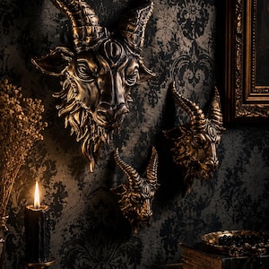 May include: Three wall-mounted goat head sculptures finished in a metallic bronze tone. Each goat head features curved horns, detailed facial features, and a textured beard. The sculptures are arranged in a cluster on a dark patterned wall.