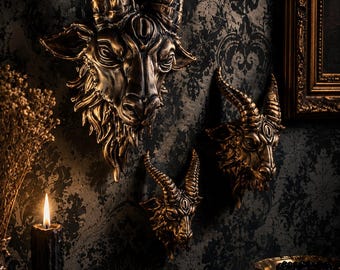 May include: Three wall-mounted goat head sculptures finished in a metallic bronze tone. Each goat head features curved horns, detailed facial features, and a textured beard. The sculptures are arranged in a cluster on a dark patterned wall.