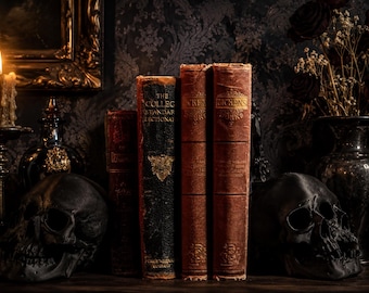 May include: A collection of four antique books, including The College Standard Dictionary and two volumes of Dickens, positioned between two black human skull replicas. A lit candle in a metal holder and a dark vase are visible nearby.