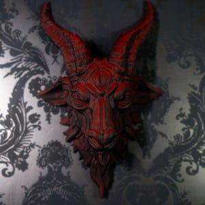 May include: A wall-mounted sculpture of a goat head featuring deep red coloring with dark shading in the crevices. The goat has prominent curved horns, detailed facial features, and a textured beard, creating a striking and dimensional appearance.
