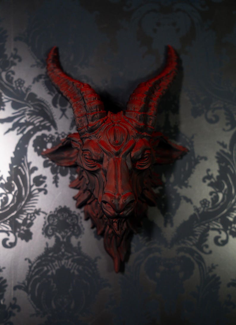 May include: A wall-mounted sculpture of a goat head featuring deep red coloring with dark shading in the crevices. The goat has prominent curved horns, detailed facial features, and a textured beard, creating a striking and dimensional appearance.