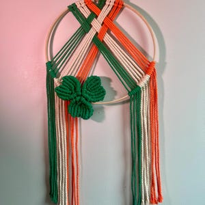 May include: A macrame wall hanging featuring a wooden hoop with green, white, and orange woven design, and a green shamrock. The colors are arranged in vertical stripes and woven patterns, creating a festive and decorative piece.
