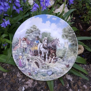 May include: A vintage ceramic plate with a colorful illustration of two horses pulling a cart across a stone bridge. The scene is set in a rural village with a church in the background. The plate is decorated with a green border and the artist's signature, "J.L. Chapman."