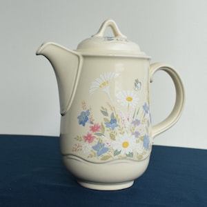 Puede incluir: Una cafetera de cerámica de color crema con un diseño floral que presenta margaritas, mariposas y otras flores silvestres en tonos azules, rosas y verdes.