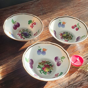 May include: Three white ceramic bowls with gold trim and fruit designs. The bowls feature a variety of fruits, including plums, grapes, berries, and apples. The bowls are on a wooden surface.