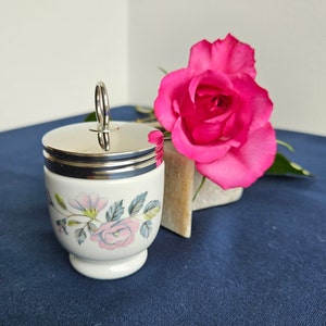 Op de afbeelding: Een wit keramiek eierdopje met een zilveren deksel en een bloemenontwerp met roze en blauwe rozen. Het eierdopje heeft een zilveren handvat en staat op een blauw oppervlak.