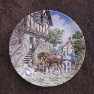 May include: A vintage ceramic plate with a colorful illustration of a horse-drawn carriage in front of a quaint cottage. The plate features a blue sky, green trees, and a stone building with a thatched roof. The plate is signed "J.L. Chapman".