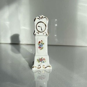 May include: A white porcelain grandfather clock figurine with gold accents and floral designs. The clock face has gold hands and Roman numerals.
