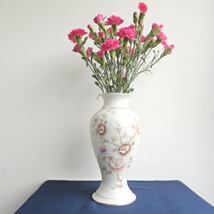 May include: A white ceramic vase with a floral pattern featuring pink roses and green leaves. The vase has a wide base and a narrow neck. It is filled with a bouquet of pink carnations.