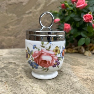 Op de afbeelding: Een wit keramiek eierdopje met een bloemenpatroon en een zilveren deksel met een ringvormig handvat. Het eierdopje staat op een stenen oppervlak.