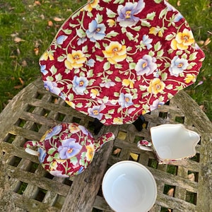 May include: A red floral tea set with a teapot, creamer, and saucer. The set features a delicate floral pattern with blue, yellow, and white flowers on a red background.