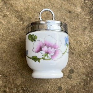 Op de afbeelding: Een wit keramiek eierdopje met een zilveren deksel en een bloemenontwerp met roze, groene en blauwe bloemen.