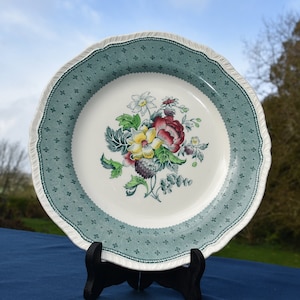 May include: A white ceramic plate with a green and white border and a floral design in the center. The flowers are red, yellow, and white.
