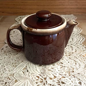 May include: A brown ceramic teapot with a white rim and a lid. The teapot has a handle and a spout. The teapot is sitting on a white lace doily.