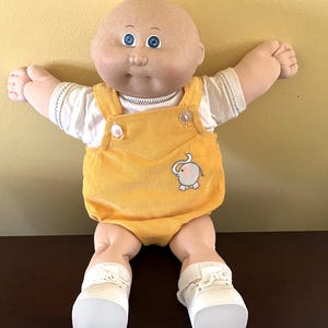 May include: A vintage baby doll with a bald head and blue eyes. The doll wears a white shirt under a yellow corduroy jumpsuit with an elephant applique. White shoes complete the outfit. The doll is sitting with arms outstretched.