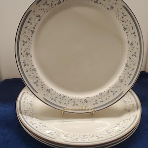 Puede incluir: Un juego de platos de cerámica blanquecina con un borde floral azul y un fino borde dorado. Los platos están apilados, con un plato ligeramente levantado, revelando el diseño. El patrón floral incluye pequeñas flores y hojas azules.