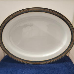 Puede incluir: Una fuente ovalada para servir con un centro blanco y un borde decorativo. El borde presenta una banda azul oscuro con un patrón dorado y marrón. La fuente está diseñada para servir alimentos.