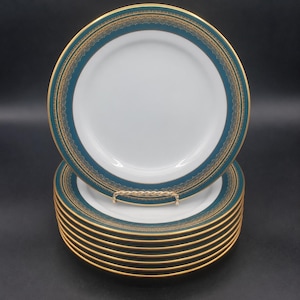 Op de afbeelding: Een stapel witte porseleinen borden met een brede blauwgroene rand versierd met ingewikkelde gouden filigraanpatronen en een dunne gouden rand. Het bovenste bord staat rechtop op een kleine standaard van goudkleurig metaal.