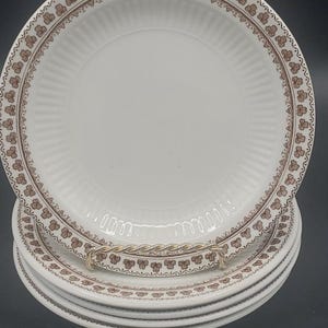 May include: A stack of white ceramic plates featuring a brown repeating clover leaf pattern along the rim. The plates have a subtle ribbed texture on the inner border and are held upright by a twisted silver-toned metal display stand.