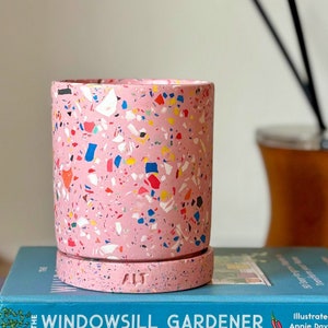 May include: Pink terrazzo planter with a white rim and a white logo that says "ALT". The planter is sitting on a blue book with the text "WINDOWSTILL GARDENER" on the cover.