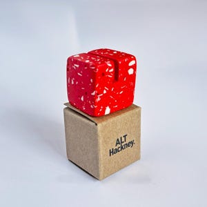 May include: A vibrant red cube, speckled with white, is slotted on top and rests on a small brown cardboard box. The box is printed with "ALT Hackney" in black. This is likely a desk accessory or a display item.