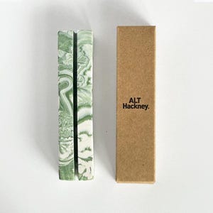 May include: A green and white marbled pen holder with a slot, next to a brown cardboard box. The pen holder has a rectangular shape and a swirled pattern. The box has the text "ALT Hackney." The design is simple and the colours are neutral.