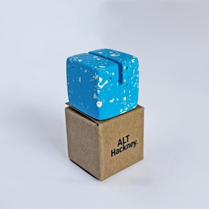 May include: A blue cube-shaped card holder with white flecks, resting on a small brown cardboard box. The box is printed with "ALT Hackney". The cube has a slot on top for holding cards or notes.