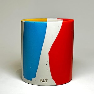 May include: A cylindrical concrete planter with a white, yellow, blue, and red abstract design. The planter has the word "ALT" printed on the bottom.
