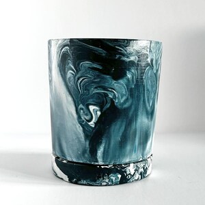 May include: A cylindrical planter with a marbled design in shades of teal, navy, and white. The swirling pattern creates a dynamic visual effect. The base of the planter has a contrasting black and white pattern.
