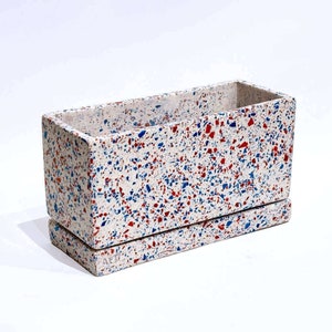 May include: A white rectangular planter with a terrazzo design featuring red, blue, and white flecks. The planter has a matching base.