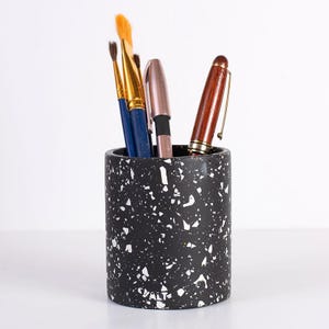 May include: A cylindrical black pen holder with a white speckled terrazzo pattern. The holder contains two paintbrushes with blue handles, a metallic pink pen, and a brown wooden fountain pen. The word CVALT is embossed near the base.