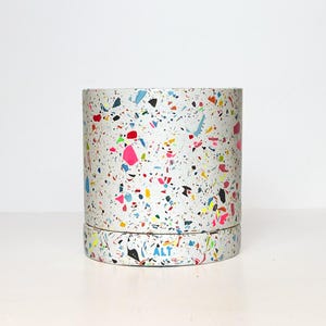 May include: A cylindrical container with a white background and a colourful terrazzo pattern. The pattern includes pink, blue, yellow, and black flecks. The word "ALT" is printed on the bottom of the container.