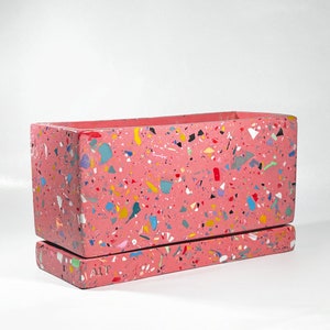 May include: A pink rectangular planter with a matching tray. The planter and tray are made of terrazzo with a variety of colourful flecks.