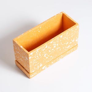 May include: A rectangular planter, approximately 20 cm long, in a warm yellow colour with a speckled white pattern. It has a matching tray at the base. The design is modern and minimalist, suitable for indoor or outdoor use. The planter is made of a solid material.