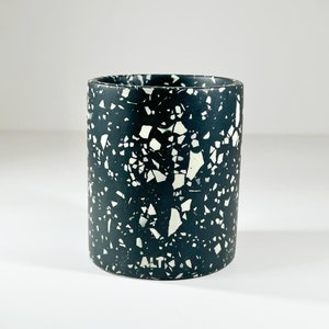 May include: A cylindrical container with a dark teal background and a speckled pattern of white and off-white fragments. The container has a smooth, matte finish and the text "ALT" is visible near the base.