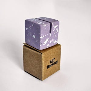 May include: A lilac cube-shaped desk accessory with a slot on top, speckled with white and gray fragments. It sits on a small brown cardboard box with the text "ALT Hackney". Likely for holding notes or photos. Measurements are approximately 5 cm x 5 cm.